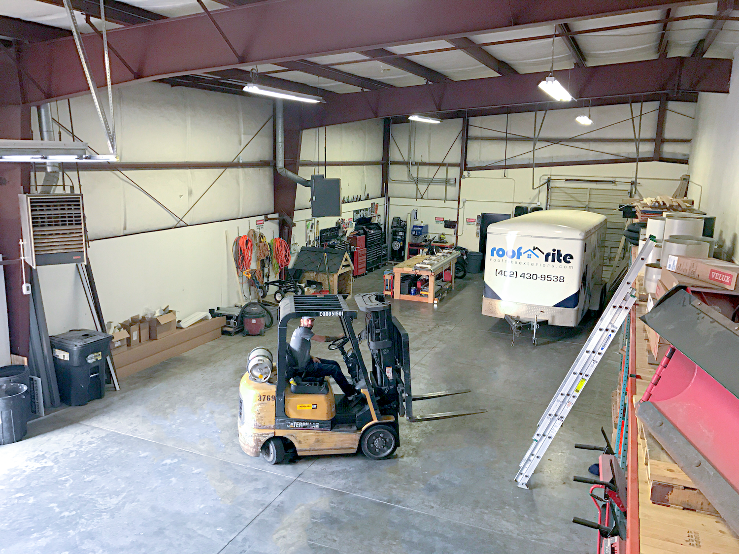 Interior view of the Roof Rite warehouse with a worker operating a forklift. A branded Roof Rite trailer is parked in the back, surrounded by tools, equipment, and roofing materials. The space is organized, with ladders, boxes , and supplies neatly stored along the walls.